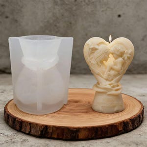 Puede incluir: Una vela encendida de color marfil en forma de corazón, con dos figuras y motivos florales, sobre una rodaja de madera. Junto a ella, un molde de silicona transparente con la forma de la vela. Se puede leer "Happy Birthday".