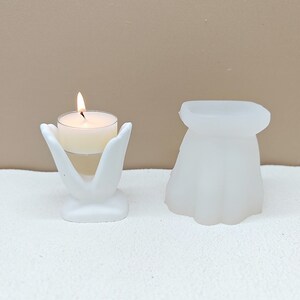 Può includere: Portacandele bianco in ceramica a forma di due mani che tengono una candela tea light. La candela è accesa e il portacandele è su una superficie bianca. C'è un secondo portacandele bianco in ceramica sullo sfondo.