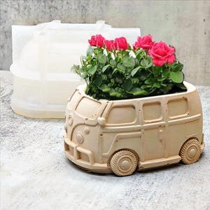 May include: A beige ceramic planter shaped like a vintage Volkswagen van with a flower pot on top. The planter is filled with green plants and pink flowers.