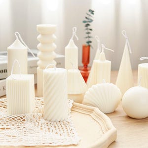 Può includere: Una collezione di candele bianche a forma di pilastro di diverse forme e dimensioni, tra cui una conchiglia, una sfera, un cono e una spirale. Le candele sono disposte su una superficie di legno con un pizzo bianco.