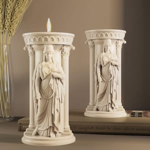May include: Two off-white candle holders in a classical column design. Each column features a sculpted figure and a round top. One candle holder has a lit candle. The candle holders are on a table.
