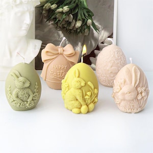 May include: Four decorative Easter egg candles. Two candles are yellow with a bunny rabbit design and flowers. One candle is green with a bunny rabbit design and flowers. One candle is beige with a floral design and the words "Happy Easter".