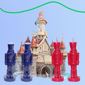 May include: Four nutcracker-shaped candles in the image. Two are navy blue, and two are red. The candles are in front of a castle with a blue sky background. The candles are standing on a round base.
