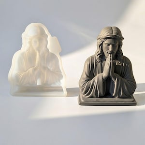 May include: A gray statue of a praying figure with long hair and a beard, hands clasped in prayer. Beside it, a translucent mold of the same figure. The statue is on a white surface.