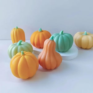 May include: A collection of six small, hand-painted ceramic pumpkins in various shades of orange, green, and yellow. The pumpkins are arranged on a white surface.