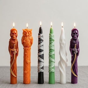 May include: Six Halloween-themed candles with lit wicks. The candles are in various shapes, including a grim reaper, an owl, a mummy, and a ghost. Colors include orange, black, green, white, and purple. Each candle has intricate details and a unique design.