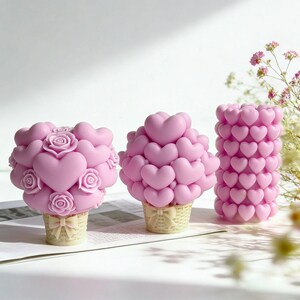 Può includere: Tre candele rosa a forma di cuori e rose. Due candele hanno la forma di bouquet in cestini con fiocchi. La terza candela è un cilindro alto ricoperto di cuori. Le candele sono su una superficie bianca.