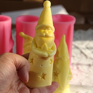 May include: A pale yellow wax figure shaped like a gnome with a tall pointed hat and a long beard, holding a star. The figure features cut-out star shapes on its body. In the background, there are several bright pink cylindrical silicone molds.