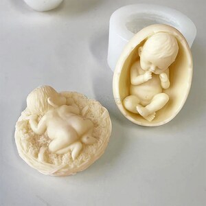 May include: Two silicone moulds for making baby figurines. One mould is a round nest with a baby lying on its stomach. The other mould is an egg shape with a baby curled up inside.