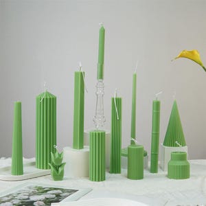 Peut inclure: Une collection de bougies piliers vertes de différentes tailles et formes. Les bougies sont disposées sur une surface blanche et certaines sont dans des bougeoirs en verre.