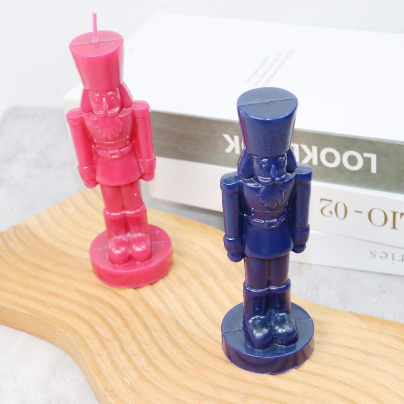 May include: Two decorative candles shaped like nutcrackers. One is a vibrant magenta, the other a deep navy blue. Both stand on circular bases and feature detailed soldier uniforms. The candles are placed on a wooden surface.