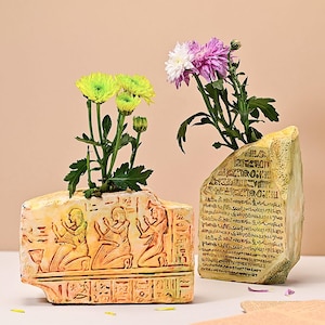 May include: Two decorative vases with an ancient Egyptian theme. One vase is rectangular with figures and hieroglyphs, the other is a stone-like shape with hieroglyphs. Both vases hold colorful flowers, creating a striking contrast.