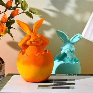 May include: Two ceramic Easter bunny figurines, one orange and one blue, each peeking out of an egg-shaped planter.