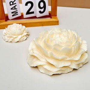 Puede incluir: Dos esculturas de flores de cerámica blanca. La flor más grande es una sola flor con múltiples capas de pétalos. La flor más pequeña es una sola flor con un diseño más compacto.