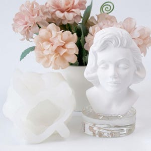 May include: A white bust of a woman with closed eyes, on a clear, circular base. A white, flower-shaped mold is to the left. Soft pink flowers in a white vase are in the background. The bust is a decorative item.