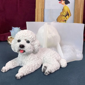 Puede incluir: Figura de cerámica blanca de un perro pequeño acostado. El perro tiene un pelaje blanco y esponjoso y la lengua rosa que sobresale. La figura está sobre una superficie gris.