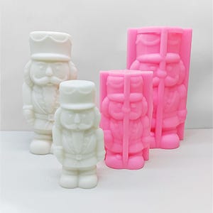 May include: Three white and pink plastic moulds of nutcracker figurines. The moulds are different sizes. The largest mould is pink and the two smaller moulds are white.