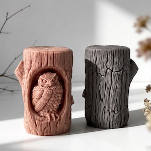 May include: Two decorative tree trunk candles. One is a light brown color with an owl carved into the front. The other is a dark gray color. Both candles have a textured, wood-like appearance.
