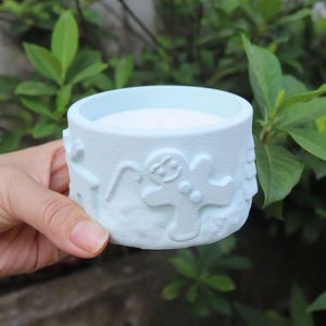 Puede incluir: Una vela azul claro en un recipiente de cerámica con un hombre de jengibre y otros diseños navideños en relieve en el lateral.
