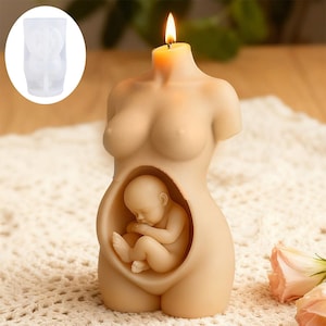 Puede incluir: Una vela beige con forma de torso de mujer embarazada con un bebé en su interior. La vela tiene una mecha encendida en la parte superior. Un molde de plástico transparente de la vela está en la esquina superior izquierda. La vela está sobre una superficie de ganchillo blanca.