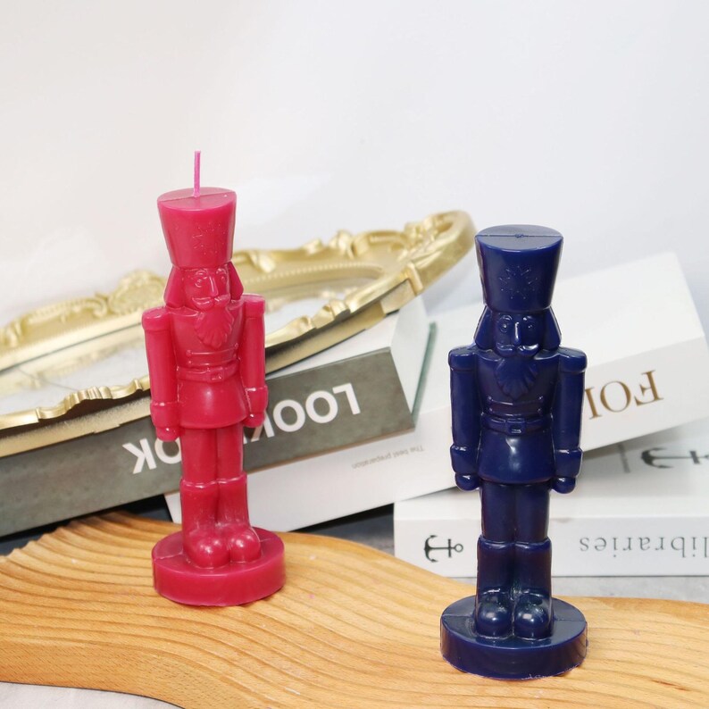 May include: Two nutcracker-shaped candles. One is a deep red color, and the other is a dark blue. Both candles have a detailed design, including a hat, uniform, and base. The candles are placed on a wooden surface.