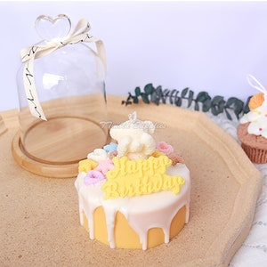 May include: A yellow and white birthday cake candle with a white frosting drip and colorful sprinkles. The candle has the words "Happy Birthday" written on it. The candle is displayed on a wooden tray with a glass dome.