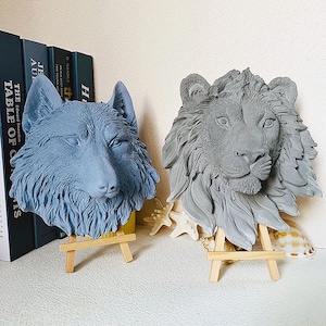 Puede incluir: Dos esculturas de yeso gris de cabezas de animales sobre caballetes de madera. La escultura de la izquierda es un lobo, y la escultura de la derecha es un león.