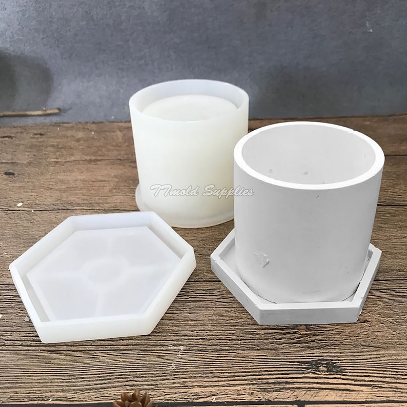 May include: Two white silicone molds for making concrete planters. The molds are in the shape of a cylinder and a hexagon. The molds are on a wooden surface.