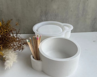 May include: White ceramic matchstick holder with a round bowl and a separate compartment for matches. The holder is sitting on a white surface with a bouquet of dried flowers in the background.