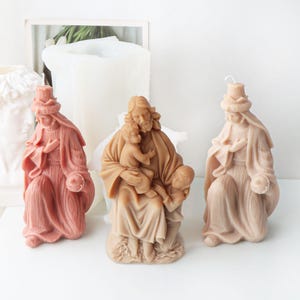 Puede incluir: Tres velas de temática religiosa en tonos rosa y beige. La vela central representa una figura que sostiene a dos niños. Las otras dos velas son de una figura vestida con una túnica. Todas las velas tienen una apariencia esculpida y detallada.
