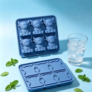 May include: Two blue silicone ice cube trays featuring six cavities each shaped like a Highland cow. One tray is filled with moulded ice figures, while the other is empty. A clear glass filled with water and ice cubes sits to the right.