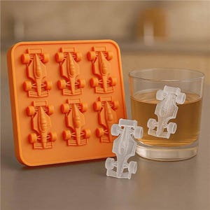 May include: Orange silicone ice cube tray with six race car molds. Two clear ice race cars are outside the tray, and two are in a glass of amber liquid. The tray is next to a glass of amber liquid.