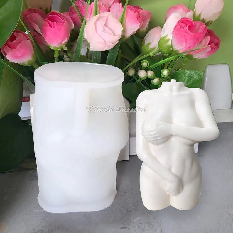 Op de afbeelding: Twee witte siliconen mallen, een in de vorm van een menselijk torso en de andere in de vorm van een cilinder. De torso mal heeft de armen gekruist over de borst. De mallen staan op een grijs oppervlak.