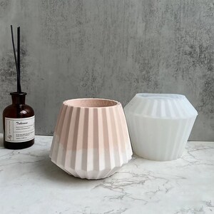May include: Two decorative geometric vases, one in a light pink and white color scheme, and the other in white. A brown glass bottle with a label and black reeds is on the left. The vases have a ribbed design.