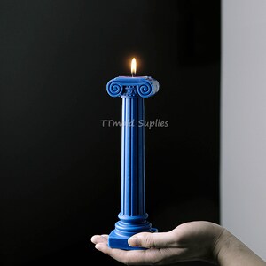 Puede incluir: Una vela azul con forma de columna romana con una llama en la parte superior. La vela est&aacute; siendo sostenida en una mano.