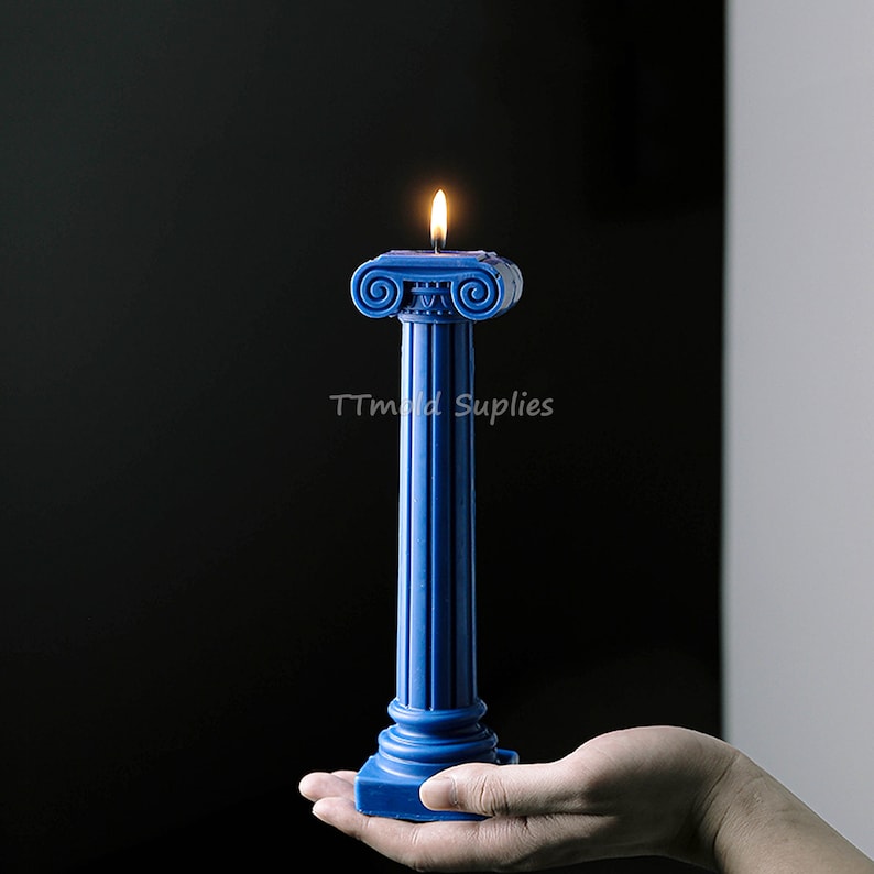 Puede incluir: Una vela azul con forma de columna romana con una llama en la parte superior. La vela est&aacute; siendo sostenida en una mano.