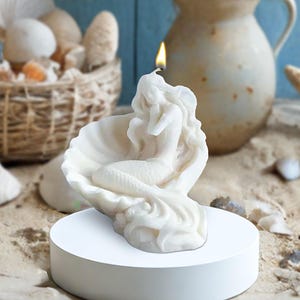 May include: White mermaid candle with a lit wick, sitting in a seashell. The candle is on a white circular base.