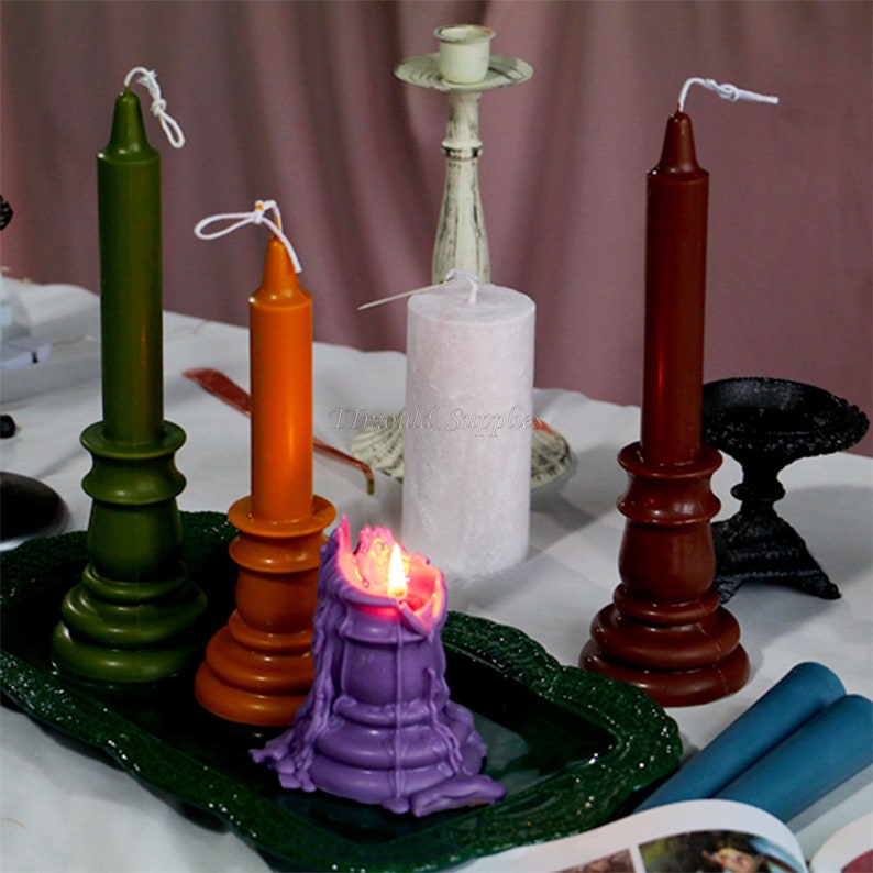 May include: Assortment of candles in various colors and styles, including green, orange, brown, purple, and white. Some candles are in decorative holders, while others are plain. The candles are arranged on a green tray with a book and a blue candle on the side.