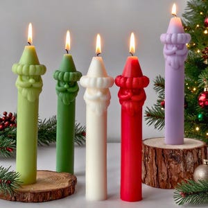 Könnte beinhalten: Fünf brennende, farbenfrohe Weihnachtsmann-Kerzen. Die Kerzen sind grün, dunkelgrün, weiß, rot und lila. Jede Kerze steht auf einer kleinen Holzscheibe. Im Hintergrund ist ein Weihnachtsbaum zu sehen.