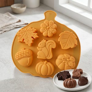 May include: A pumpkin-shaped silicone baking mold in a mustard yellow color featuring cavities for a squirrel, an acorn, two pumpkins, and two different leaf shapes. A small white plate sits in front with four dark chocolate pieces in these shapes.