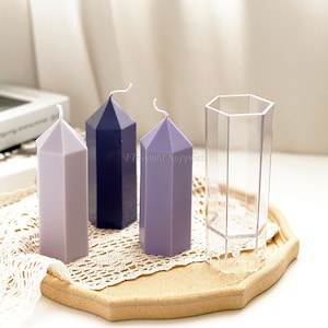 Puede incluir: Tres velas hexagonales en forma de pilar en tonos de morado y azul, con un molde hexagonal de plástico transparente. Las velas están sobre una bandeja de madera con un encaje blanco.