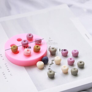 May include: A pink silicone mold with six round cavities for making small, round candies or decorations. The mold is 7 cm in diameter. There are 12 finished candies in various colors, including purple, brown, beige, and gray. Each candy is approximately 1 cm in diameter.
