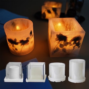 以下が含まれることがあります： 白いベースが付いた、正方形が2つと円形が2つの透明なプラスチック製のキャンドルモールド4つ。モールドは、円形が2つと正方形が1つの、花柄の3つの点灯したキャンドルの隣に置かれています。