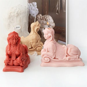 May include: Three decorative candles shaped like a sphinx. The candles are in different colors: red, pink, and beige. The candles are on a white surface. The word "Smile" is written on a small white tag.