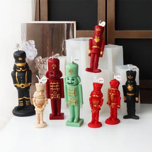 May include: A set of ten nutcracker-themed candles in various colors, including red, green, and black. The candles are all standing upright on a white surface.