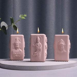 May include: Three pink rectangular candles with detailed relief carvings of Buddha figures. Each candle has a lit wick, casting a warm glow. The candles are arranged on a light gray, circular tray, with a dark gray background.