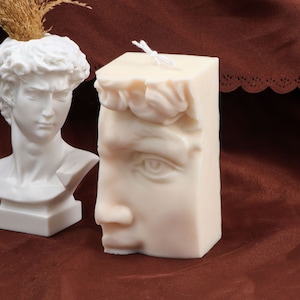 Puede incluir: Una vela blanca esculpida en forma de rostro de hombre. La vela está sobre una superficie marrón.