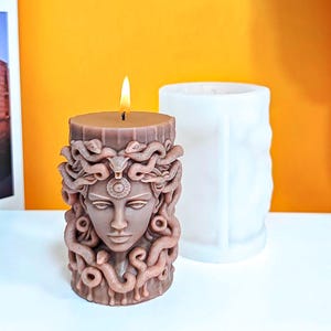 May include: A brown cylindrical candle featuring a sculpted face surrounded by snakes, with a lit wick at the top. Beside the candle sits a white silicone mold with a matching textured design, used for creating the candle shape in a cylindrical form.
