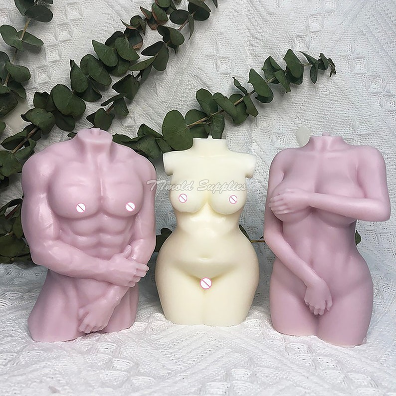 Op de afbeelding: Drie naakte torso-kaarsen, twee zijn roze en &eacute;&eacute;n is wit. De kaarsen zijn allemaal koploos en hebben armen. De kaarsen zijn in een rij op een wit oppervlak gerangschikt.
