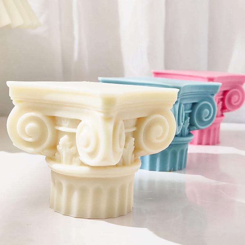 K&ouml;nnte beinhalten: Drei dekorative Kerzenhalter in Form von ionischen S&auml;ulen in Creme, Blau und Rosa. Die Kerzenhalter weisen detaillierte Schn&ouml;rkel und kannelierte S&auml;ulen auf, die eine klassische &Auml;sthetik schaffen. Die S&auml;ulen sind in einer Reihe angeordnet.